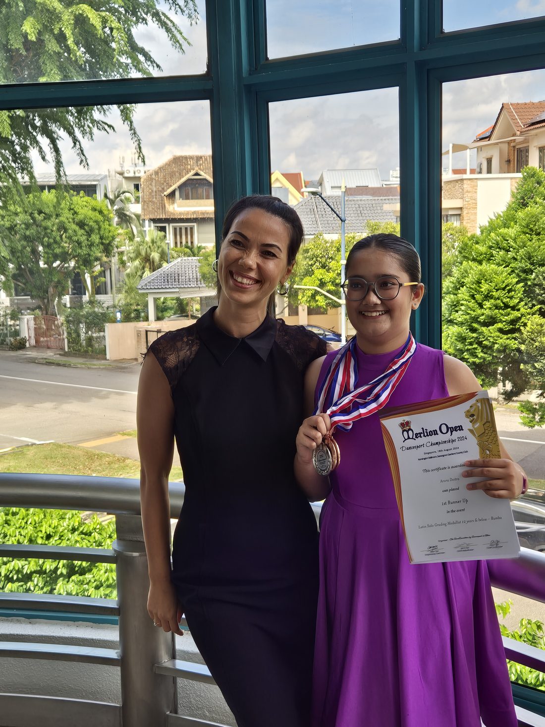 Raliza with a young student in a purple dress wearing a medal outside a competition venue