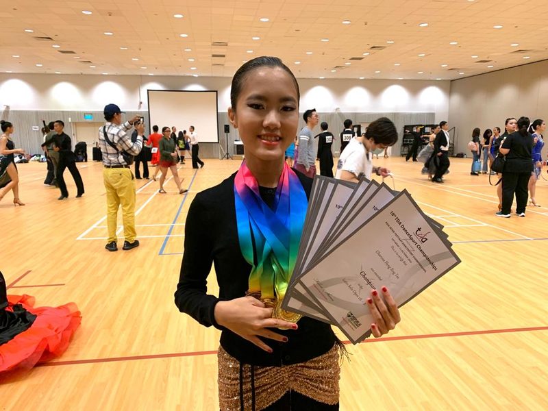 A young student in a school hall holding a stack of medals and certificates