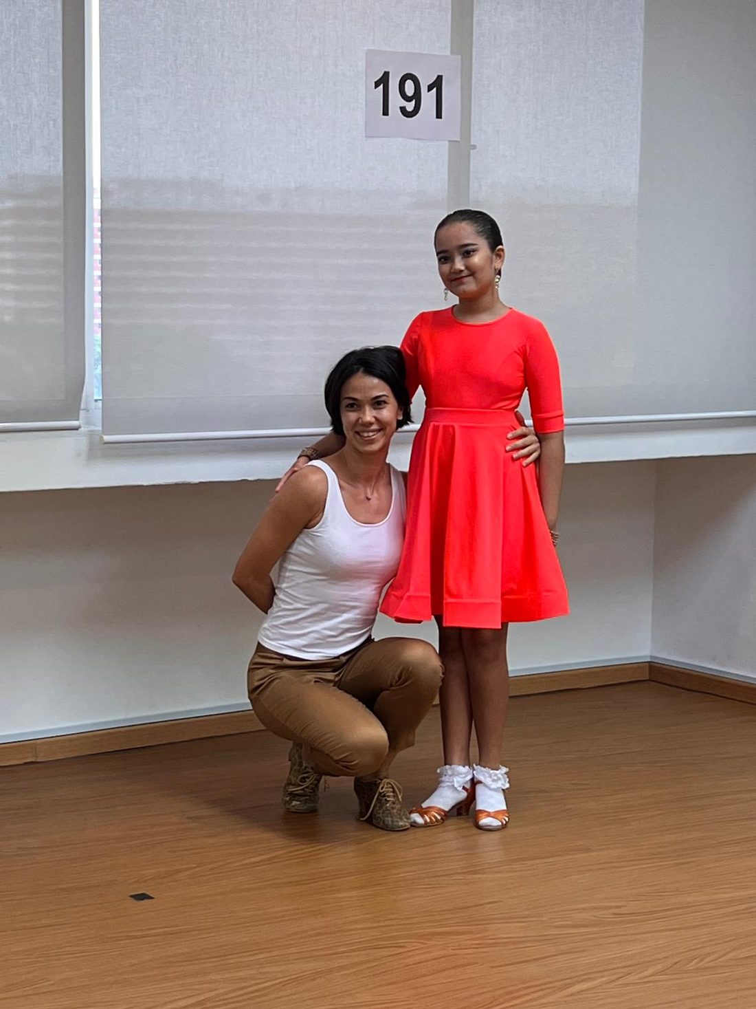 Raliza kneeling beside a young student in a red practice dress in a studio
