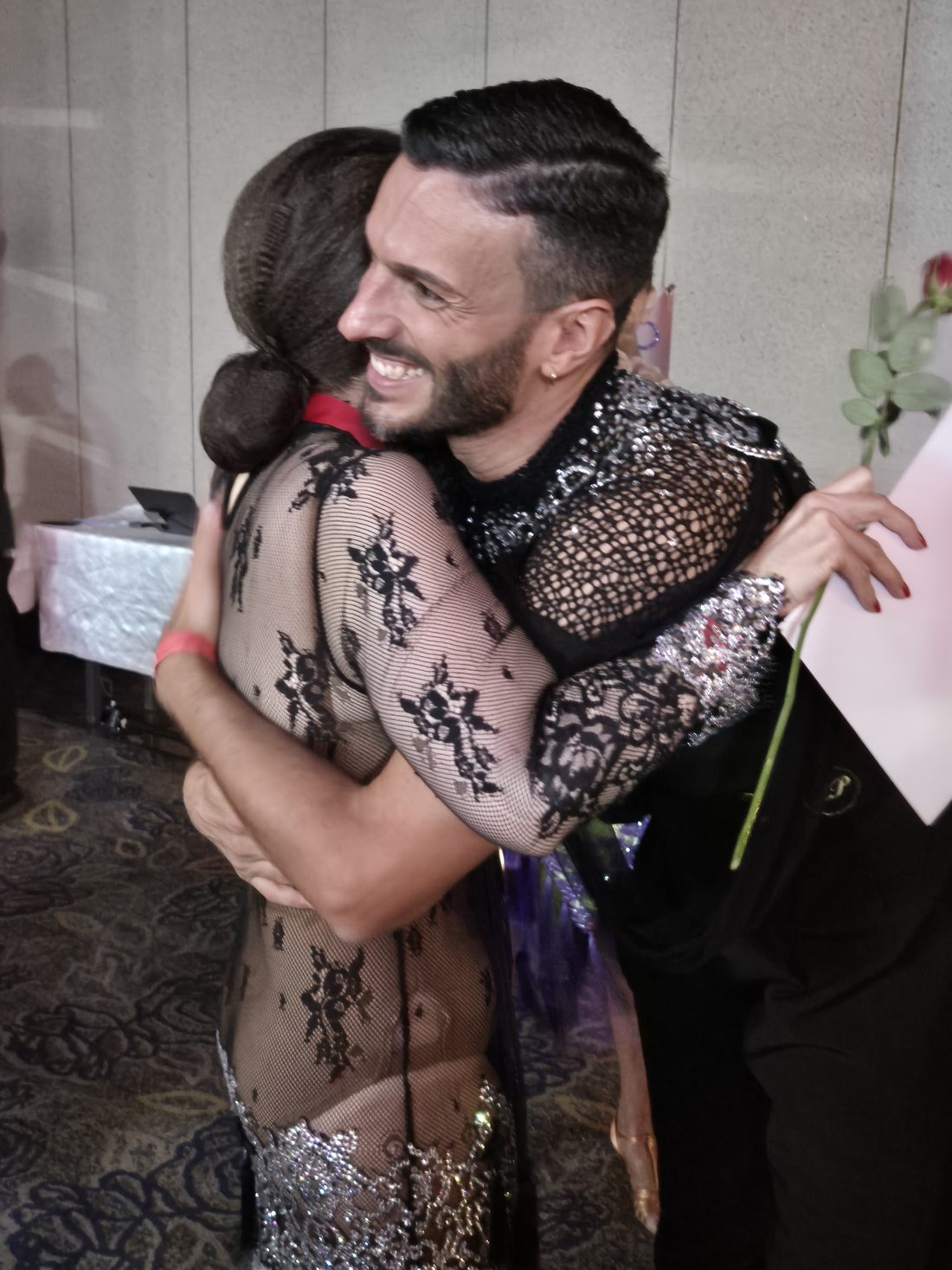 A student in a lace competition dress hugging her partner, holding a rose and a certificate