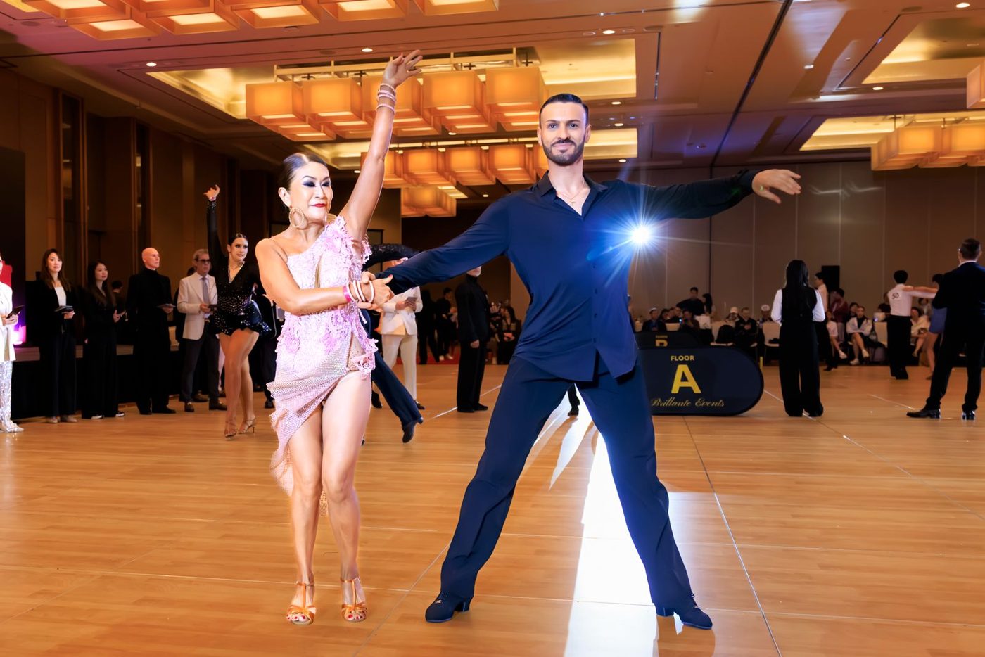 Raliza dancing with a pro-am partner in a dark suit on a crowded competition floor